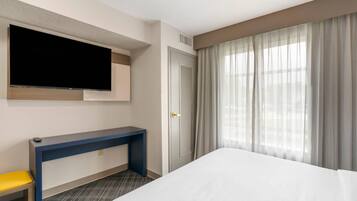 Habitación de hotel con cama, televisor montado en la pared, escritorio con silla y ventana con cortinas.
