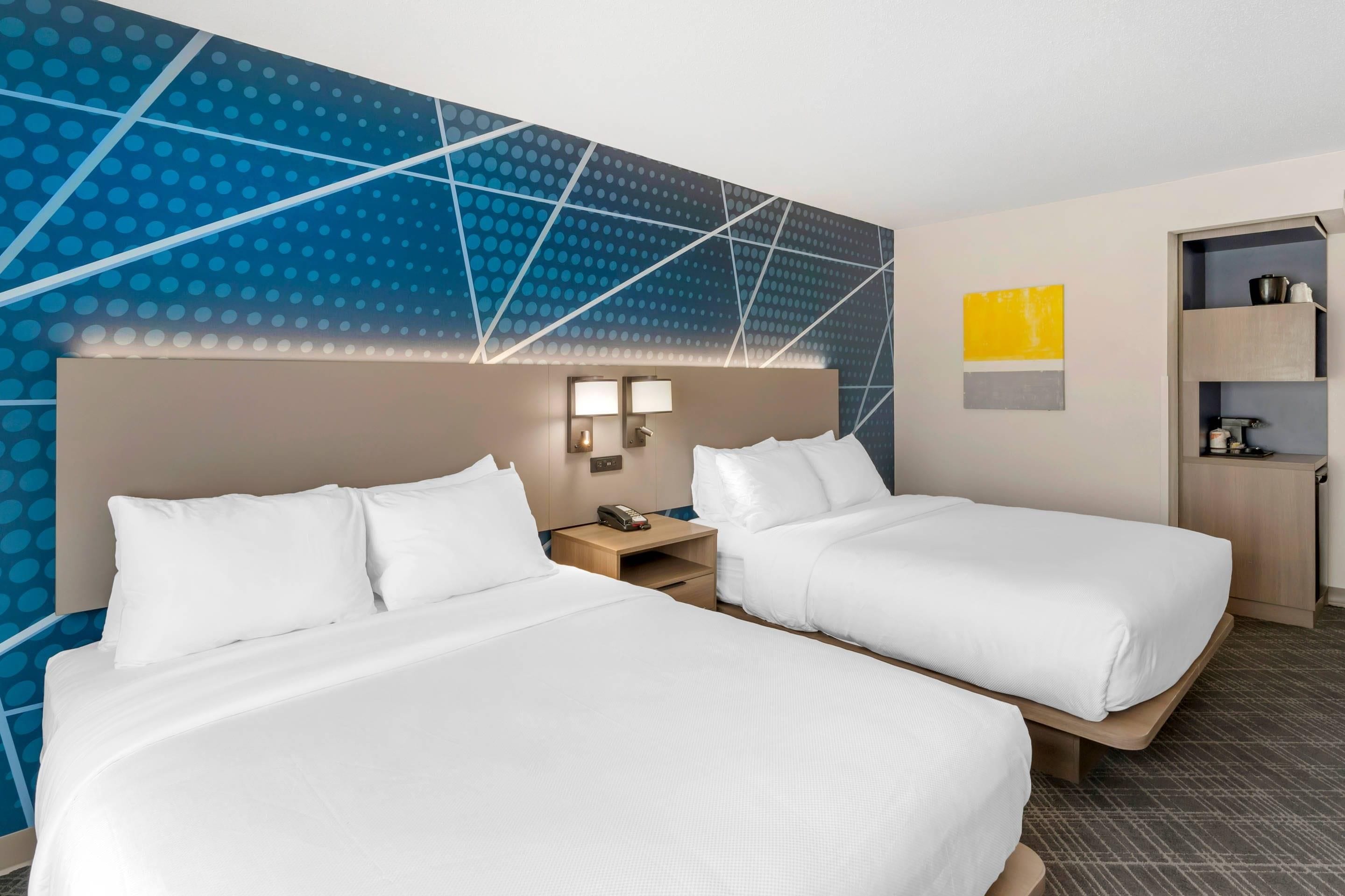 Habitación de hotel con dos camas, una mesita de noche, una lámpara de pared y un estante con un hervidor.