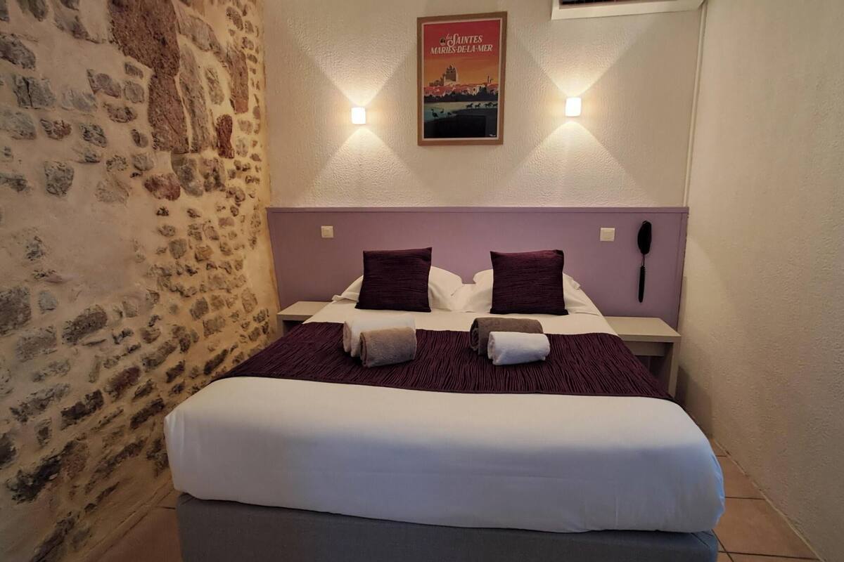 A bedroom with a stone wall, a bed with white bedding and two dark pillows, a small bedside table with a phone, and a poster on the wall.