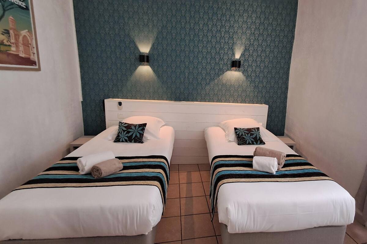 A double room with two beds, each with a striped blanket and a pillow. There are two wall-mounted lights above the beds, and the room has a tiled floor.