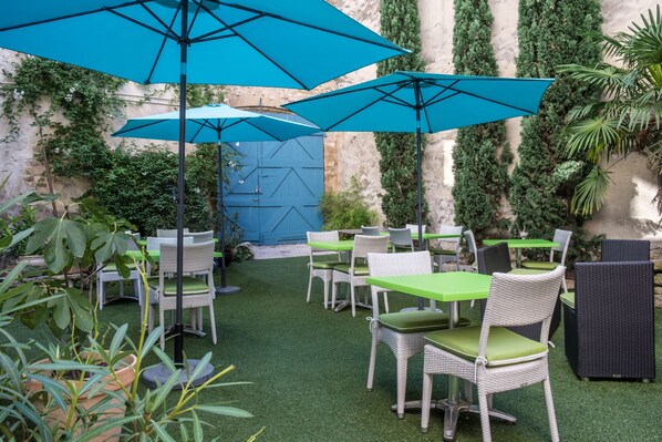 Un patio al aire libre con mesas y sillas verdes, sombrillas azules y abundante follaje.