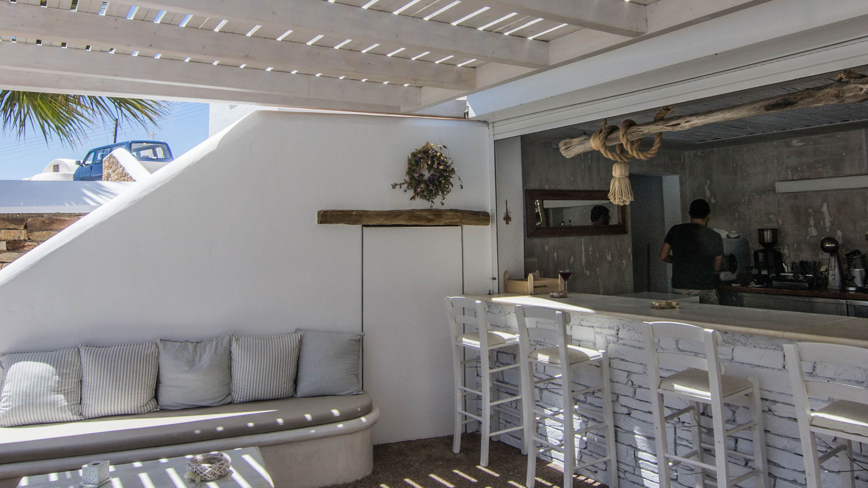 A modern outdoor seating area with a white bench and white chairs, a wooden shelf with a floral arrangement, and a bar area with a large mirror and rustic decor.