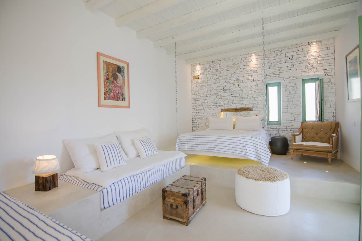Un dormitorio con una cama a rayas blancas y azules, un sofá blanco, un baúl de madera y un reposapiés blanco.