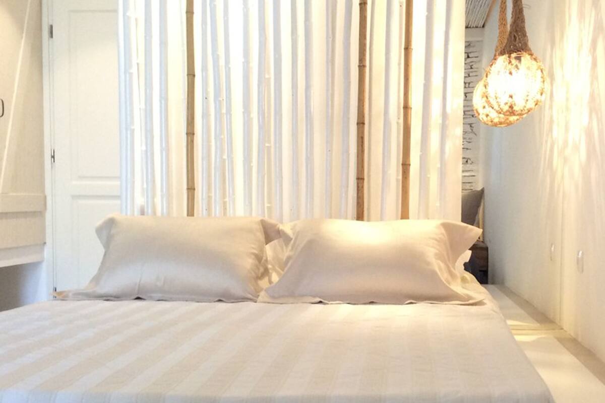 Una cama bien hecha con sábanas y almohadas blancas, un lámpara colgante y persianas verticales de bambú al fondo.