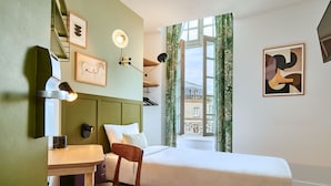 A hotel room with a bed, a desk, a chair, a window with curtains, and artwork on the walls.