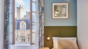 A bedroom with a bed, a nightstand, a wall-mounted lamp, a framed picture, and a window with curtains.