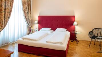 Una habitación de hotel con una cama tapizada en rojo, dos mesitas de noche con lámparas, suelo de madera y una silla.