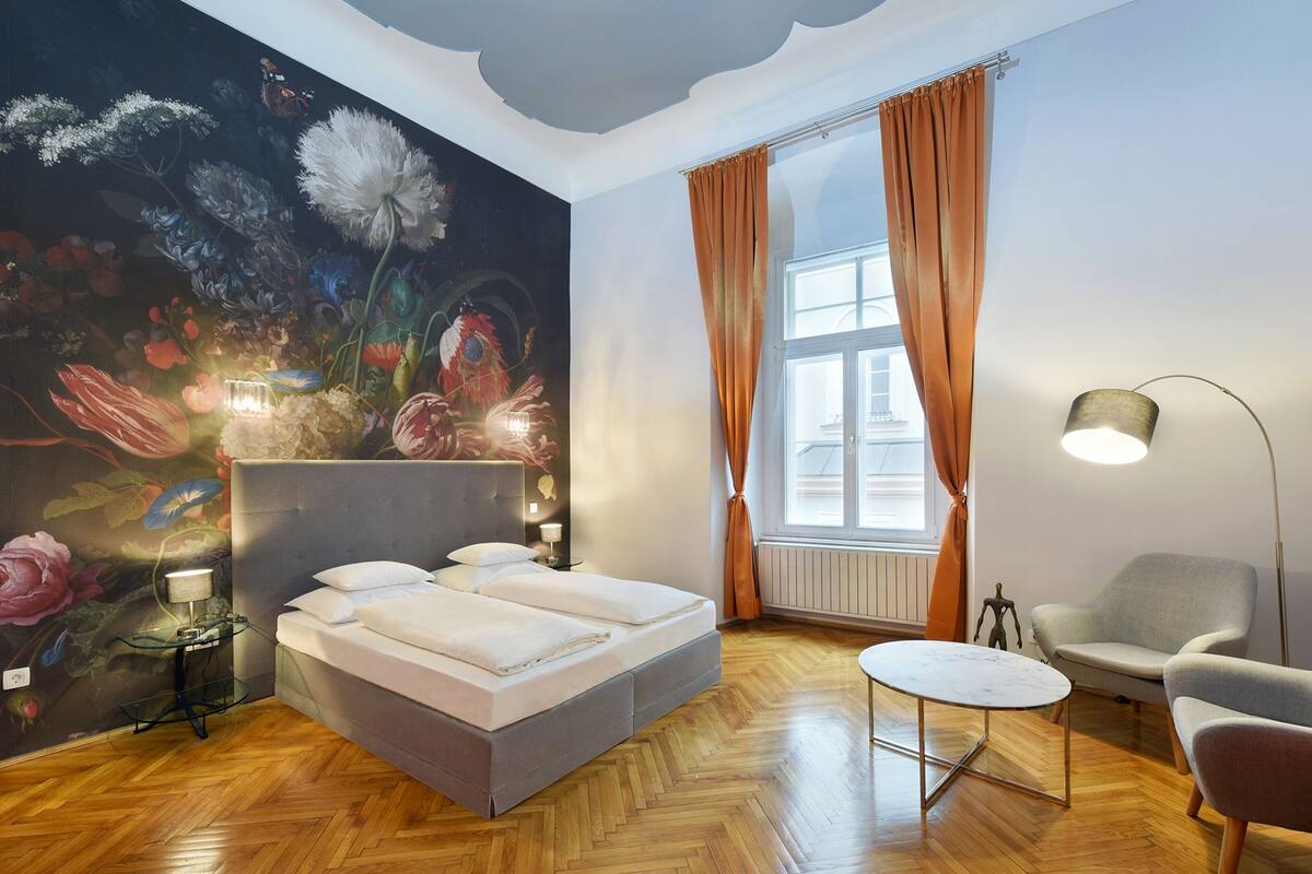 Een slaapkamer met een groot bed, een klein rond tafeltje, een stoel, een lamp en een bloemen behang.