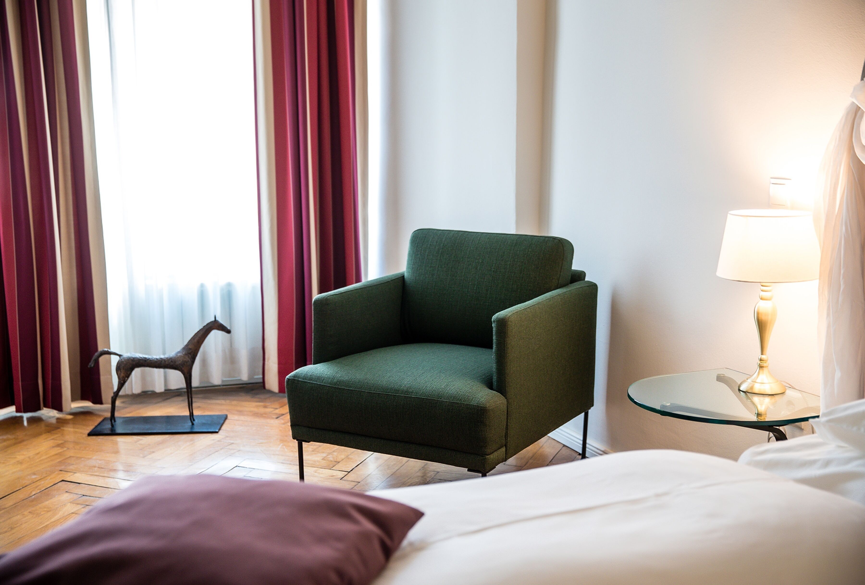 A hotel room with a green armchair, a glass side table with a lamp, a wooden floor, a bed with white bedding, and a red curtain.