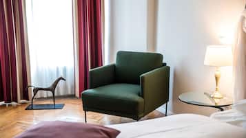 A hotel room with a green armchair, a glass side table with a lamp, a wooden floor, a bed with white bedding, and a red curtain.