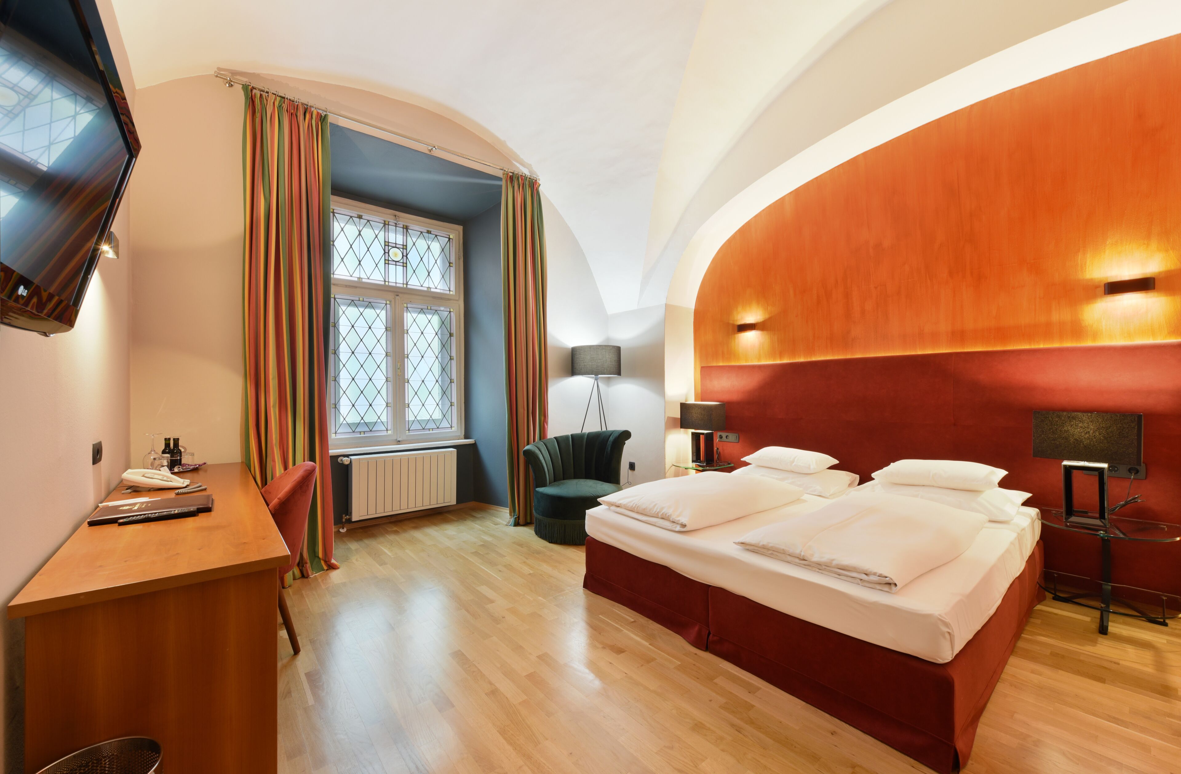 Ein Hotelzimmer mit einem Holztisch, einem Bett mit weißen Bettwäsche, einem grünen Sessel und einem an der Wand montierten Fernseher.