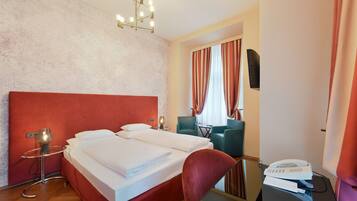 Una habitación de hotel con cabecera roja, cama con ropa blanca, una mesa de vidrio con una lámpara, una silla, un escritorio con teléfono y un televisor en la pared.