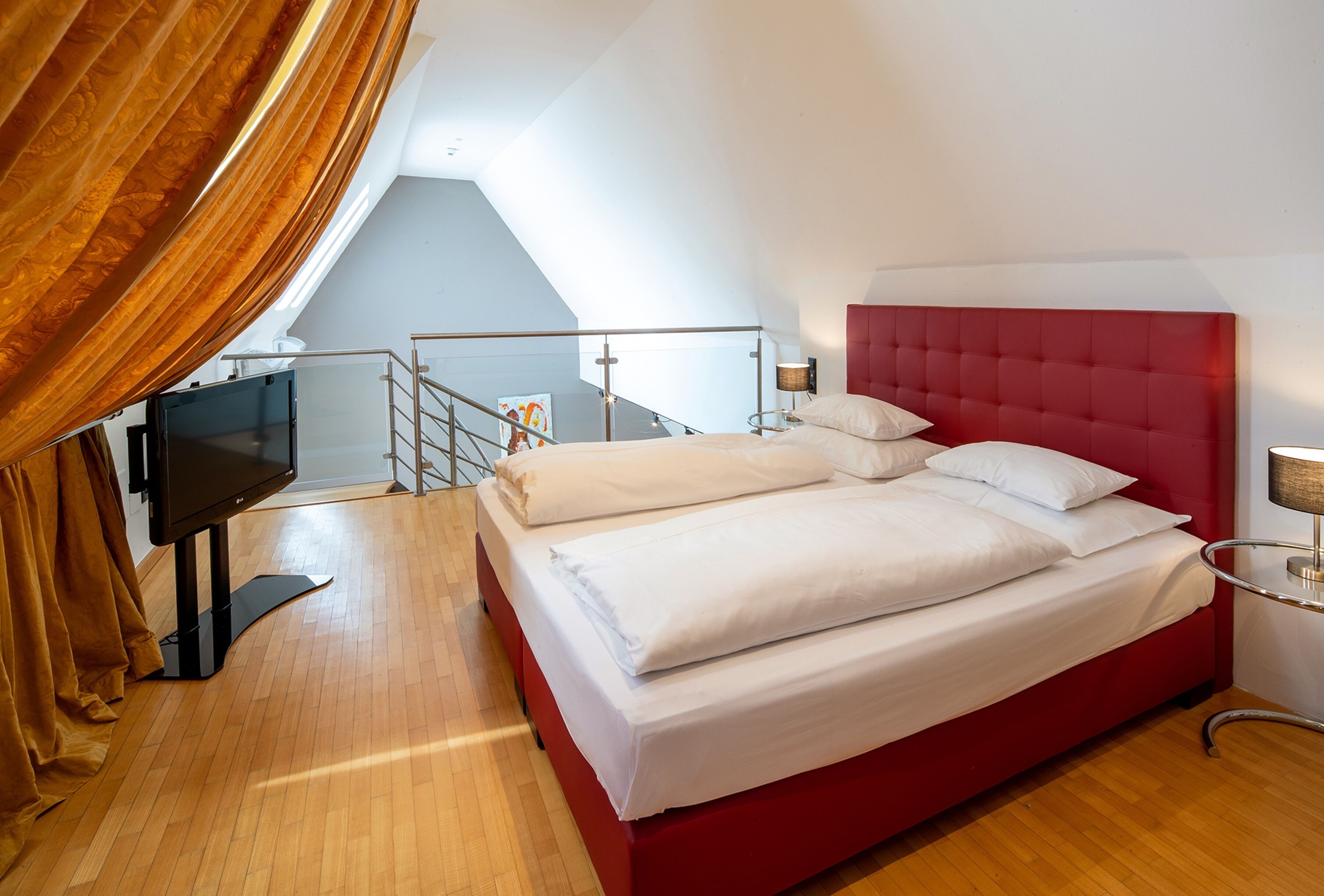 Una habitación de hotel con una cama roja, ropa de cama blanca, un televisor sobre un soporte, una escalera y vistas al cielo.