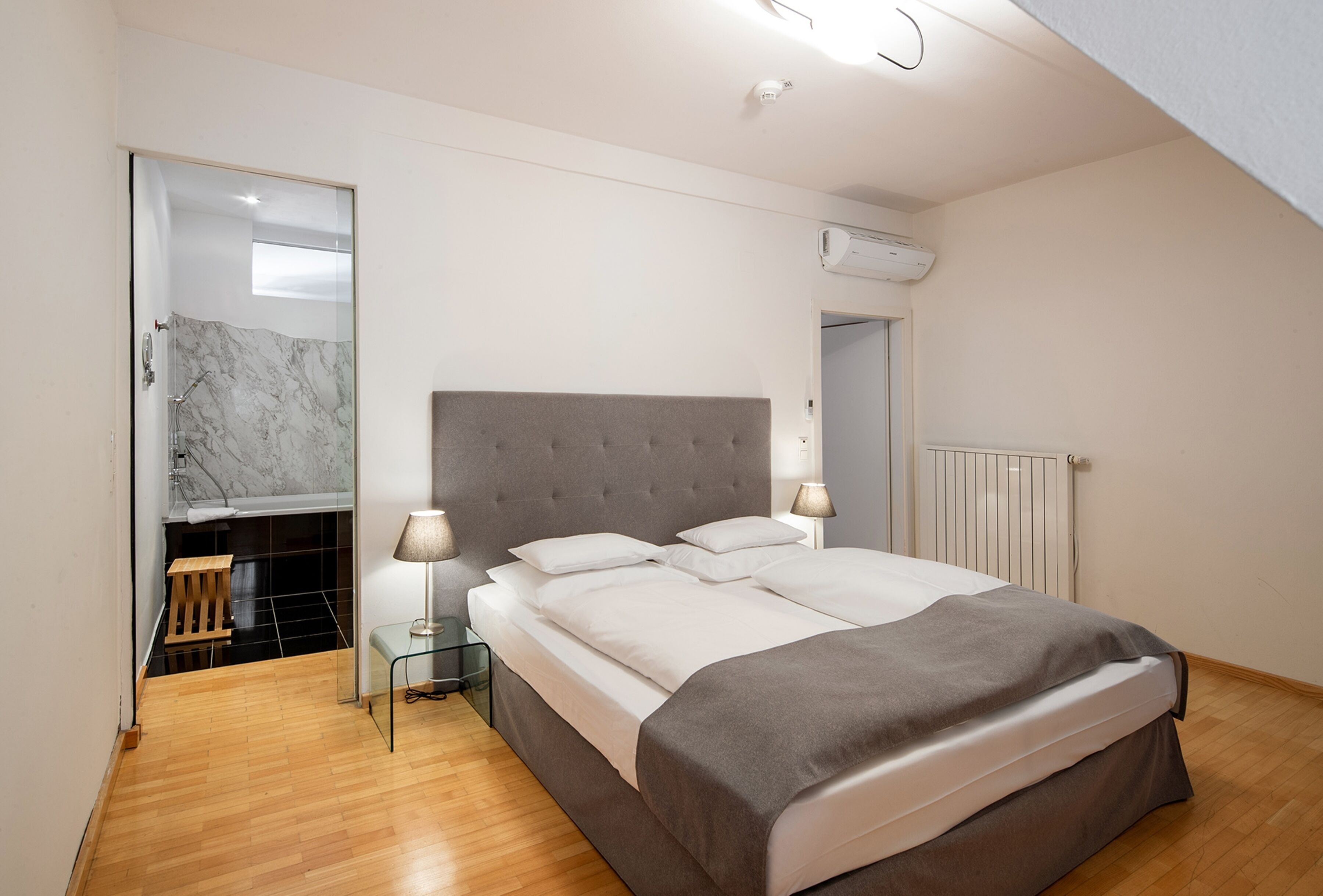 A bedroom with a large bed, a bedside table, a lamp, a mirror, and a bathroom with a shower and a marble-tiled wall.