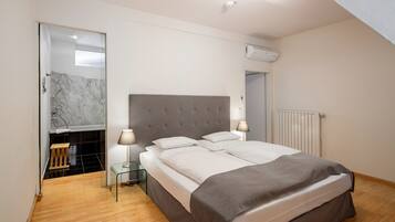 A bedroom with a large bed, a bedside table, a lamp, a mirror, and a bathroom with a shower and a marble-tiled wall.