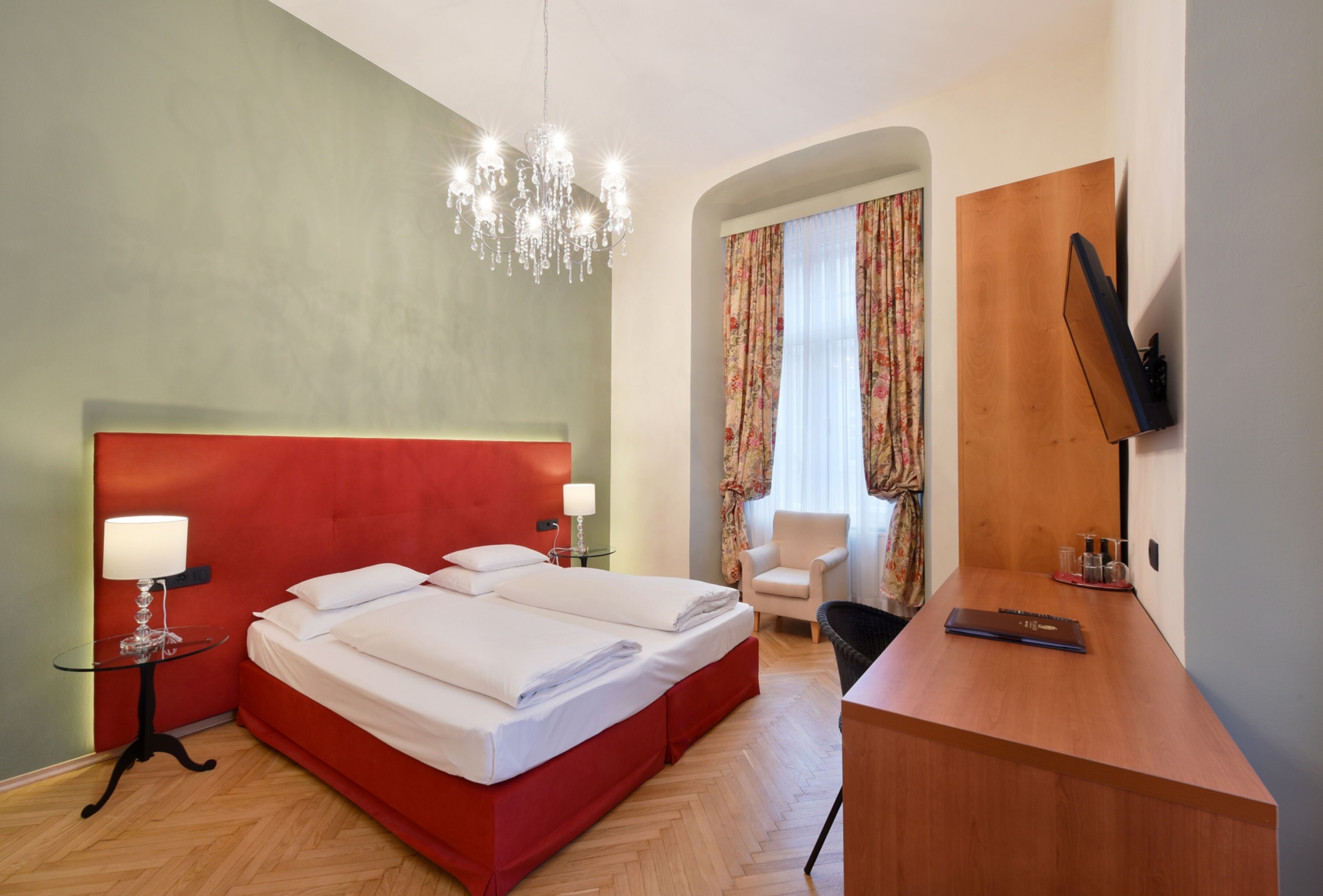 A hotel room with a red headboard, a bed with white linens, a desk with a laptop, a chair, and a TV mounted on the wall.