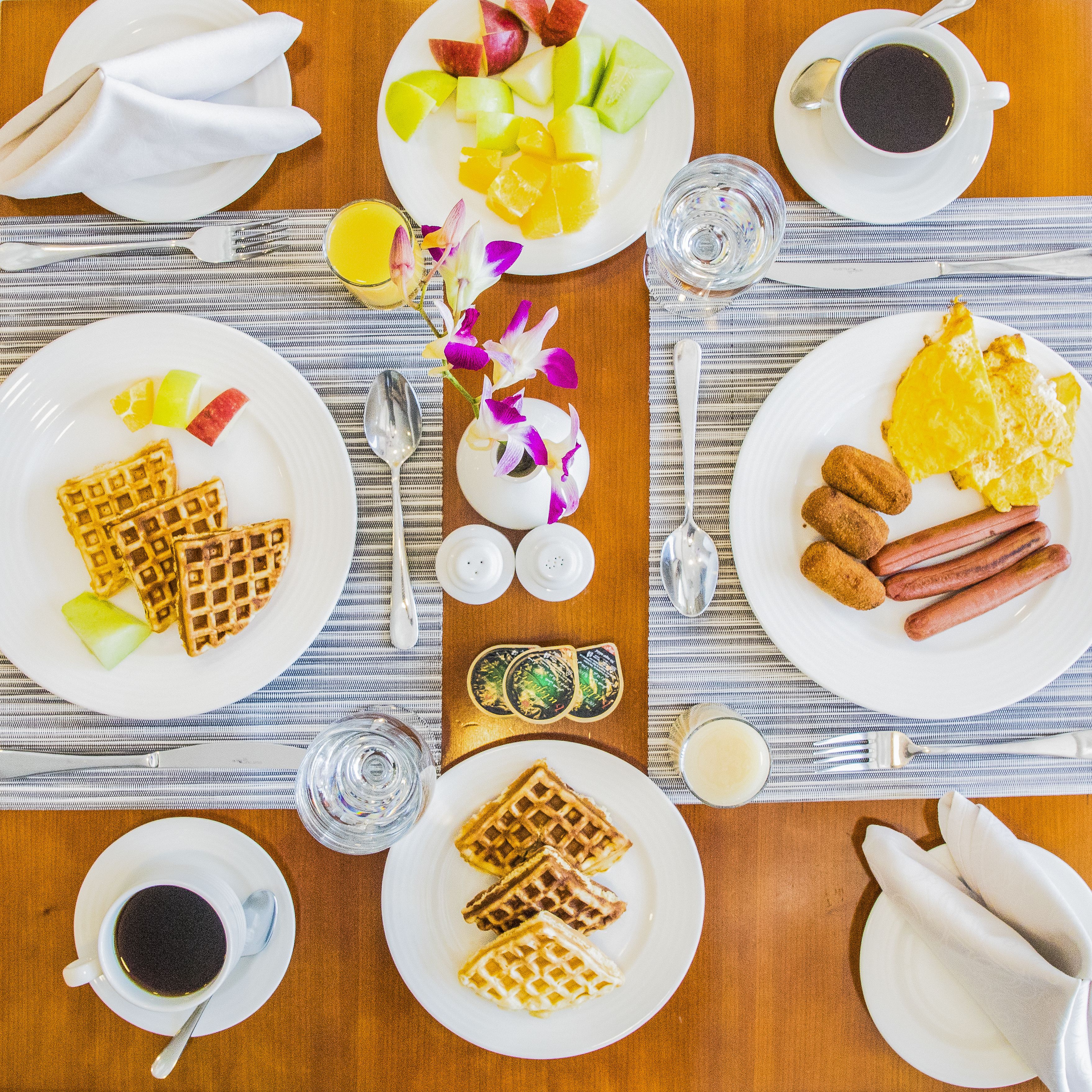 Mesa de café da manhã com waffles, frutas, cachorros-quentes e café.