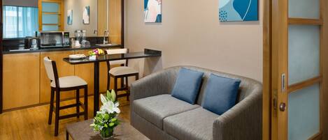 Una habitación de hotel moderna con un sofá gris, dos cojines azules, una mesa de centro de madera con un arreglo floral, un área de comedor con una mesa y sillas negras, una cocina con microondas y máquina de café, y obras de arte en las paredes.