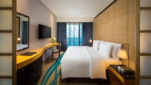 A hotel room with a large bed, a desk with a flat-screen TV, a chair, a desk lamp, and a window with curtains.