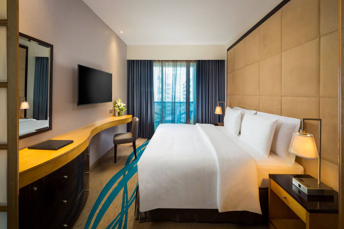 A hotel room with a large bed, a desk with a flat-screen TV, a chair, a desk lamp, and a window with curtains.