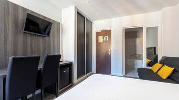 Quarto de hotel moderno com cama, escrivaninha com cadeiras, televisão de tela plana e porta deslizante para um banheiro.