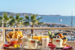 A breakfast tray with various foods and drinks, a basket of bread, and a view of the sea and palm trees.
