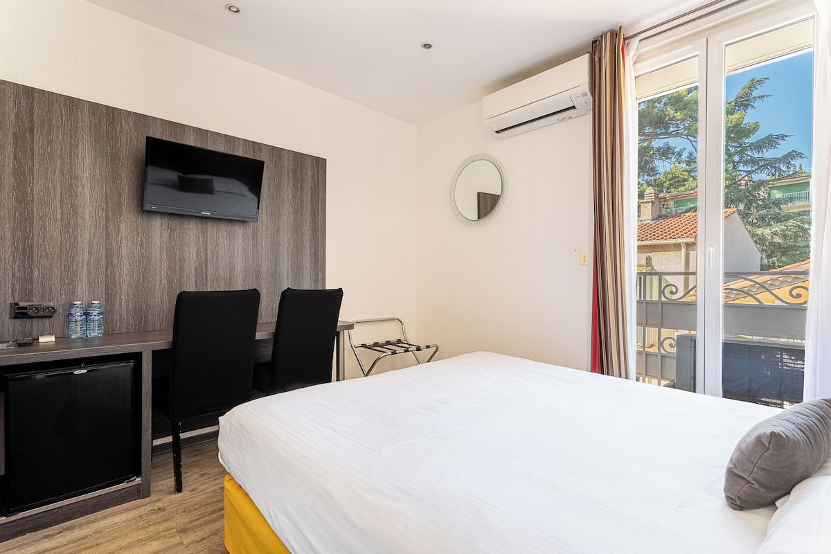 A hotel room with a bed, a desk with a TV, a chair, a small fridge, a mirror, and a balcony with a view of buildings and trees.