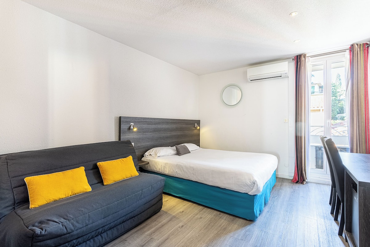 A compact hotel room with a sofa, a single bed, a small table, and a chair.