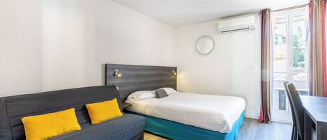 A compact hotel room with a sofa, a single bed, a small table, and a chair.