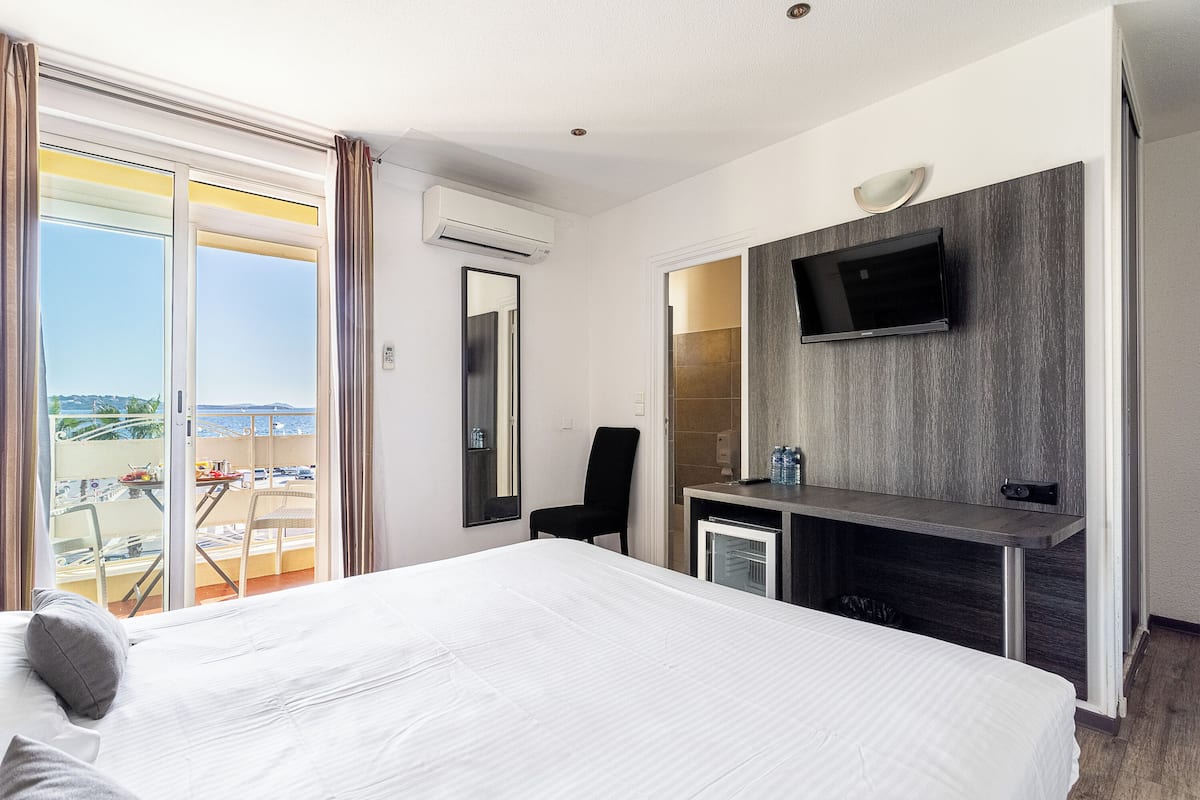A hotel room with a large bed, a desk with a TV, a chair, and a balcony with a view.