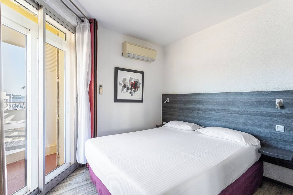 A hotel room with a bed, a picture on the wall, an air conditioning unit, and a sliding glass door to the balcony.