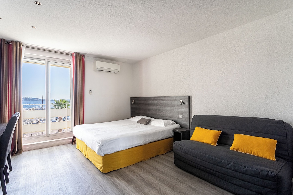 A hotel room with a bed, a sofa, a desk, and a large window with a view of the beach.