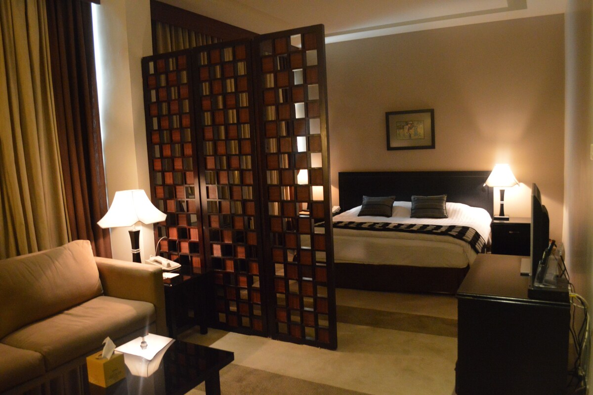 A hotel room with a bed, a sofa, a desk, a lamp, and a partition with a grid pattern.