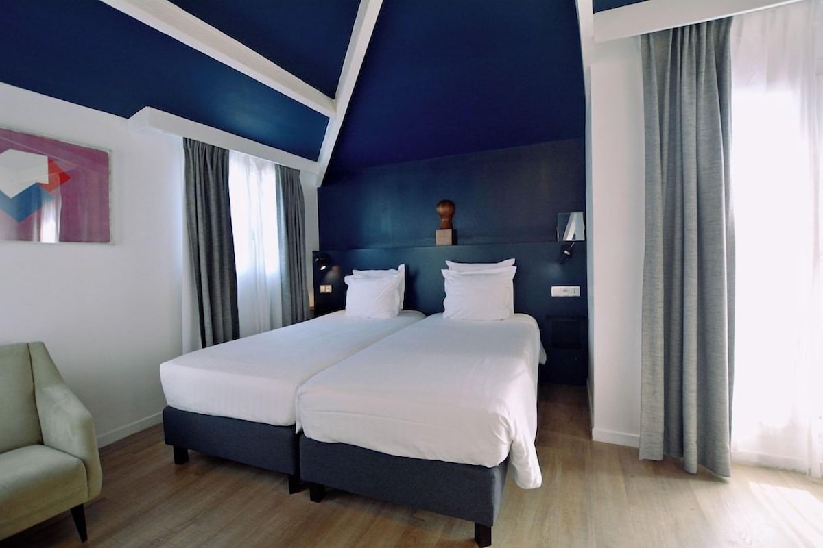 A hotel room with two beds, a chair, a desk, and a painting on the wall.