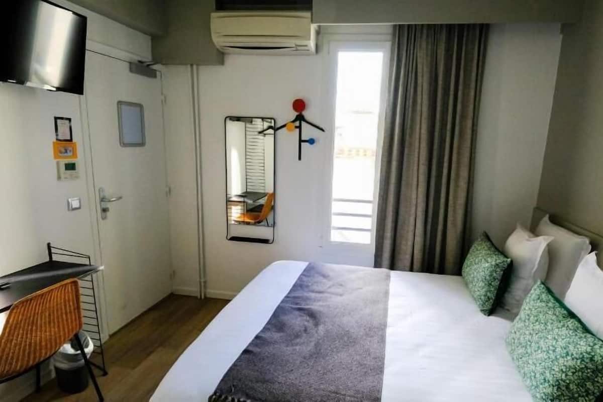 Una camera d'albergo con un letto, una scrivania, una sedia, una TV, una finestra con tende e un condizionatore d'aria a parete.