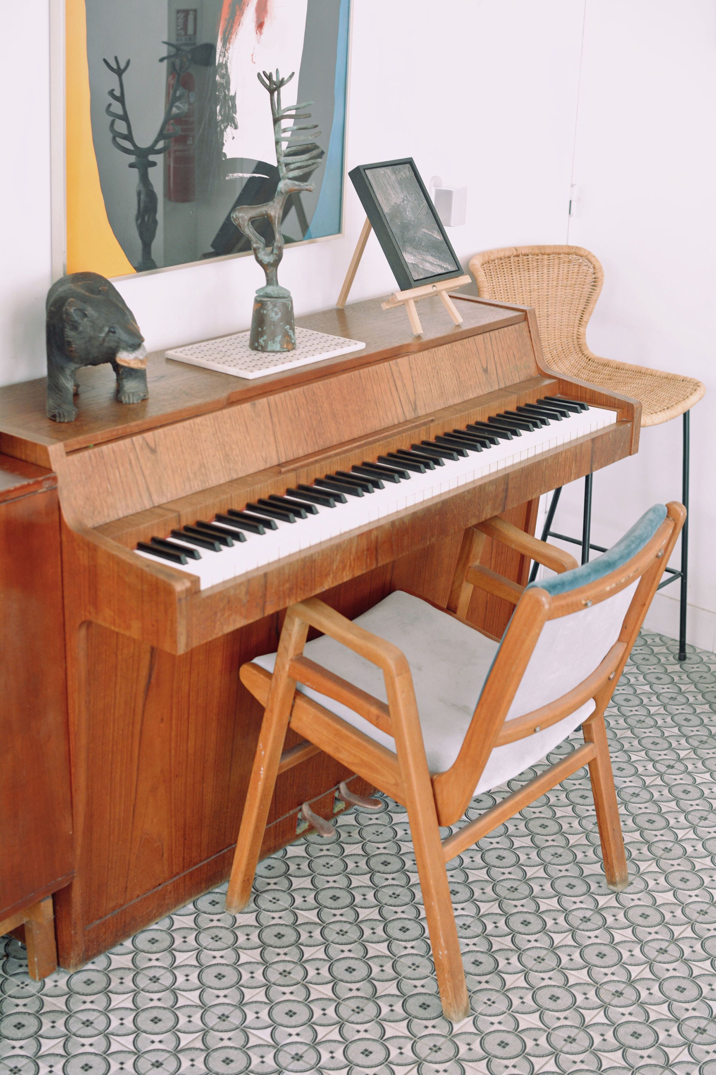 Un piano de fusta amb un tamboret, una cadira i un quadre encimbellat a dalt.