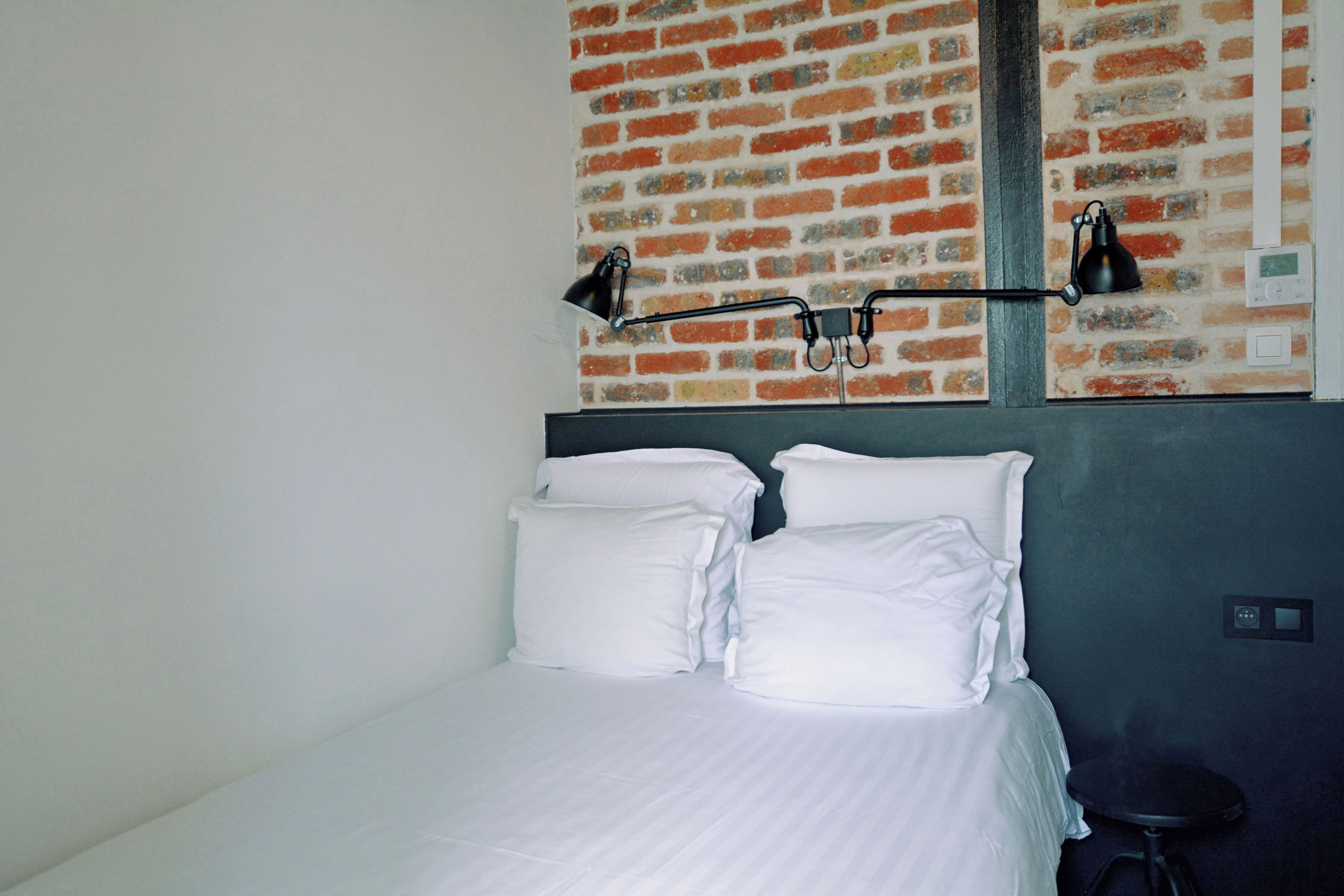 Quarto de hotel com parede de padrão de tijolos, uma cama com roupa de cama branca e duas luminárias fixadas na parede.
