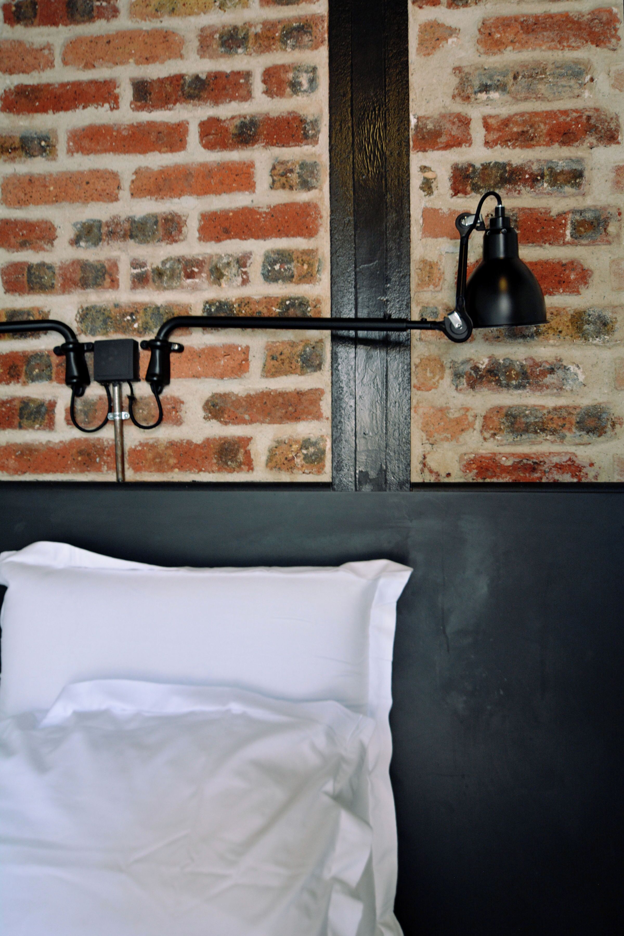 A bed with white linens, a brick wall, and a black lamp on a shelf.