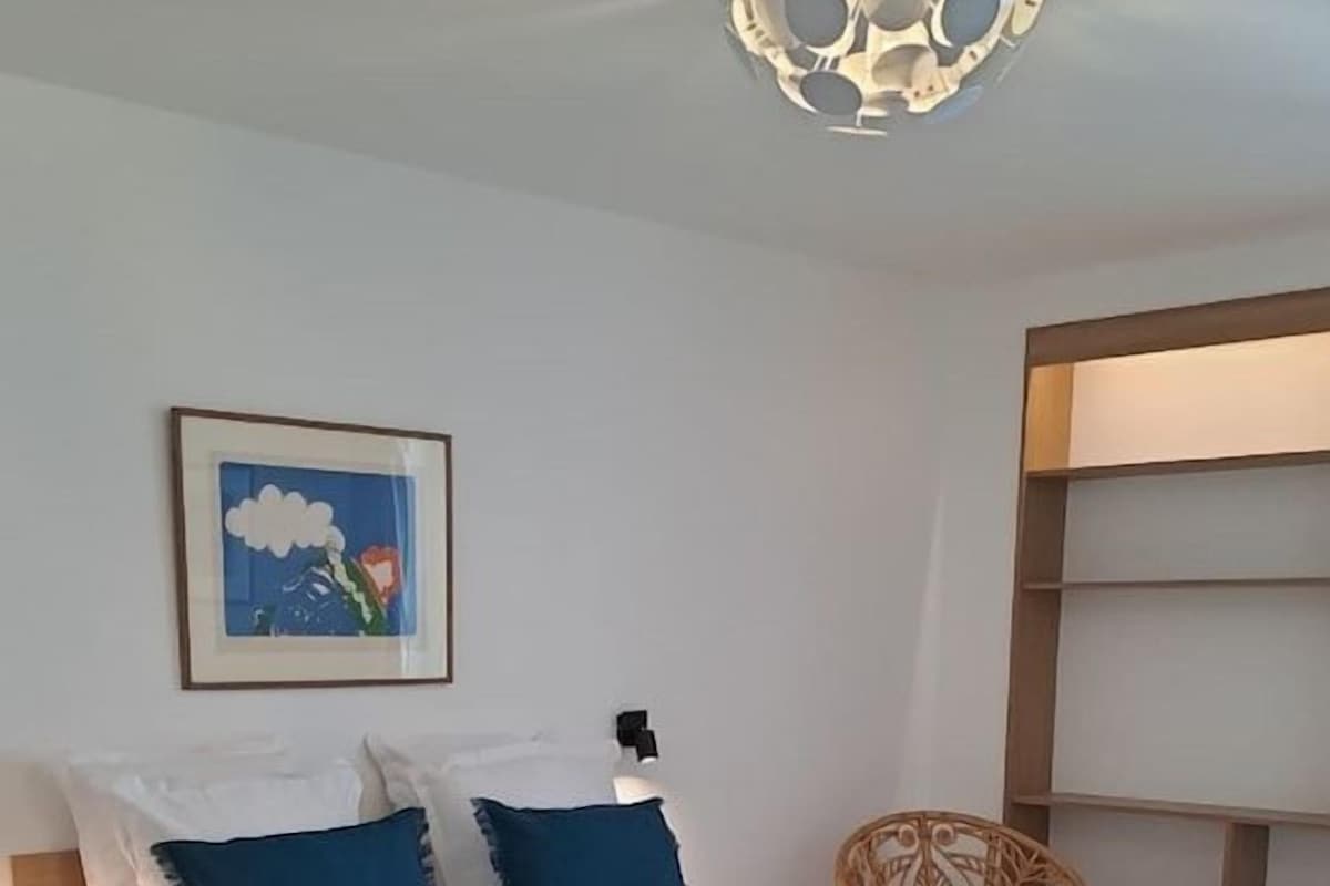 A bedroom with a bed, a chair, a bedside table, and a wall-mounted light fixture.