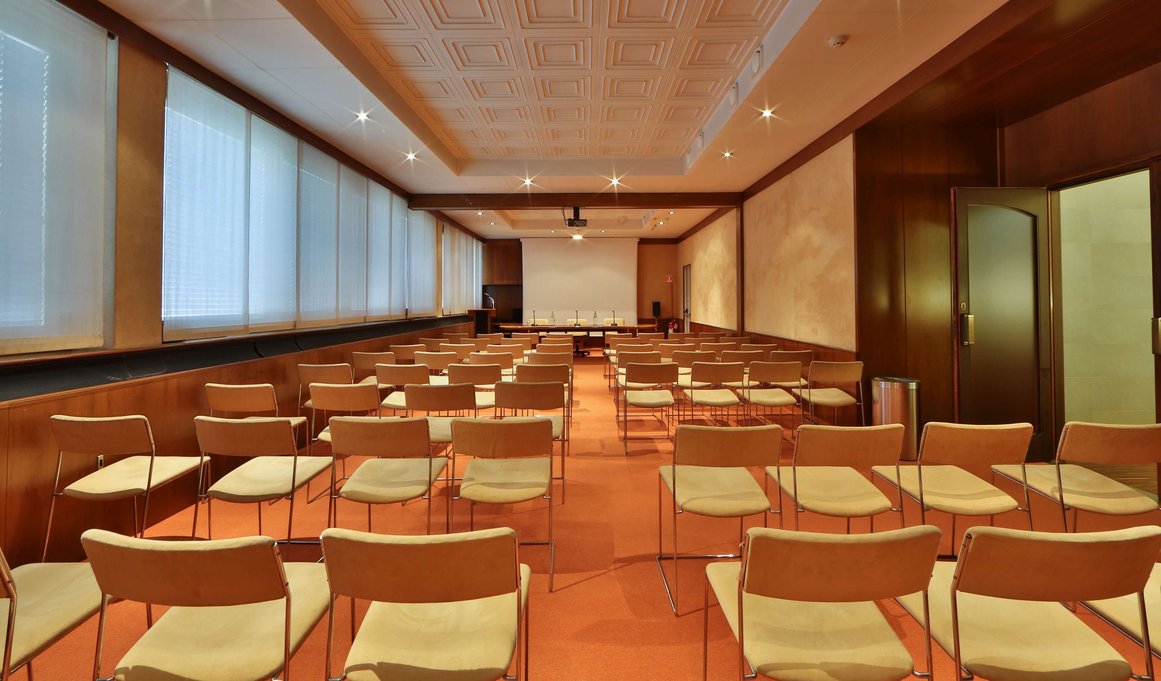 Un salón de conferencias con filas de sillas, una pantalla de proyector y un ventanal grande.