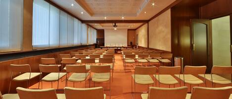 Un salón de conferencias con filas de sillas, una pantalla de proyector y un ventanal grande.