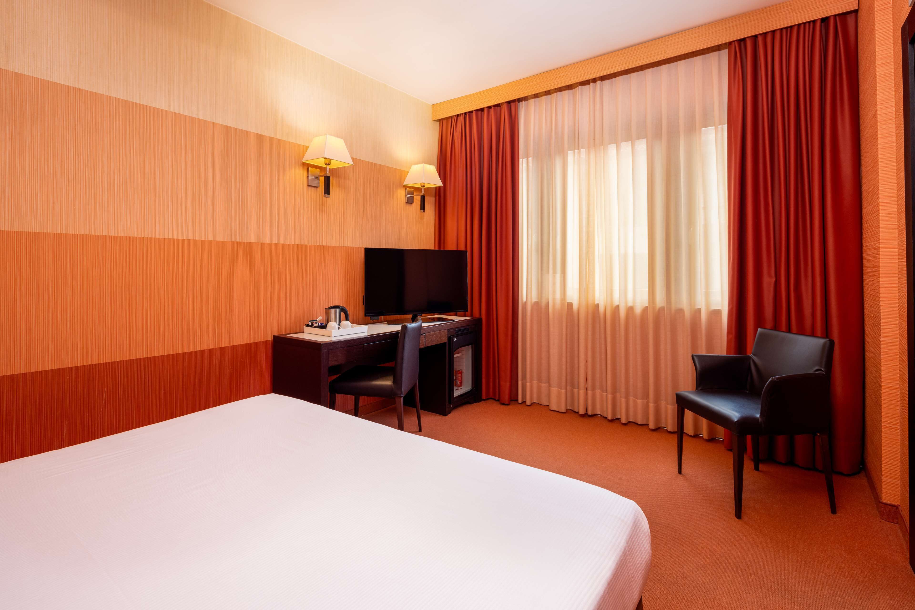 Ein Hotelzimmer mit einem großen Bett, einem Schreibtisch mit Fernseher, zwei Stühlen und einem Fenster mit Vorhängen.