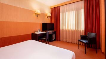 Ein Hotelzimmer mit einem großen Bett, einem Schreibtisch mit Fernseher, zwei Stühlen und einem Fenster mit Vorhängen.