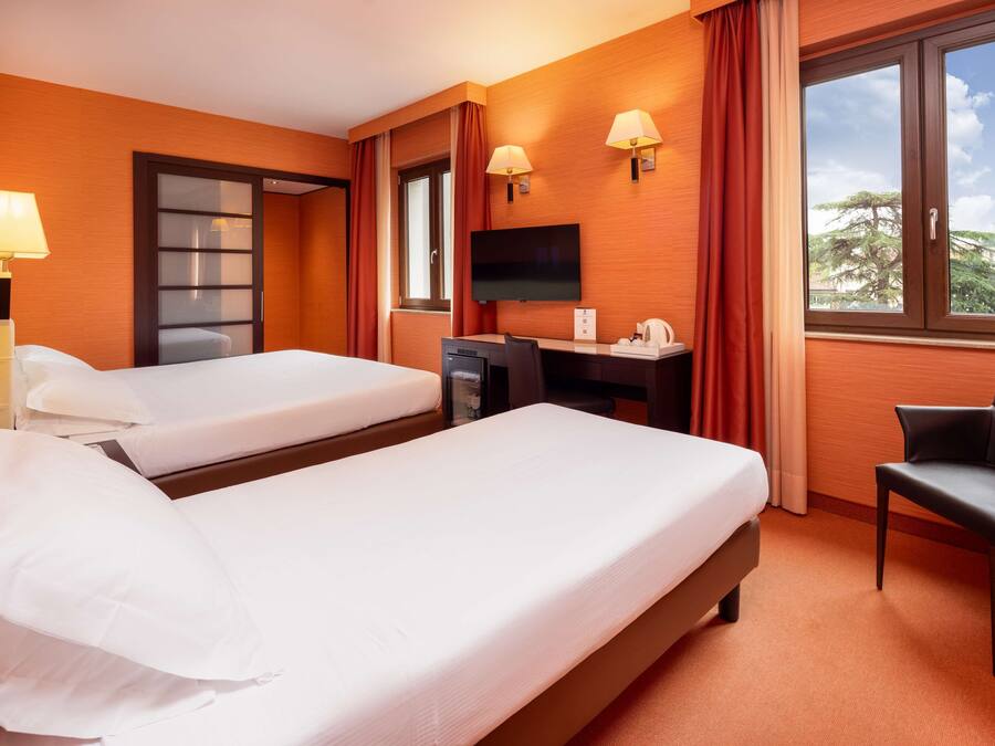 Une chambre d’hôtel avec deux lits, un bureau, une chaise, une télévision et une fenêtre donnant sur des arbres.