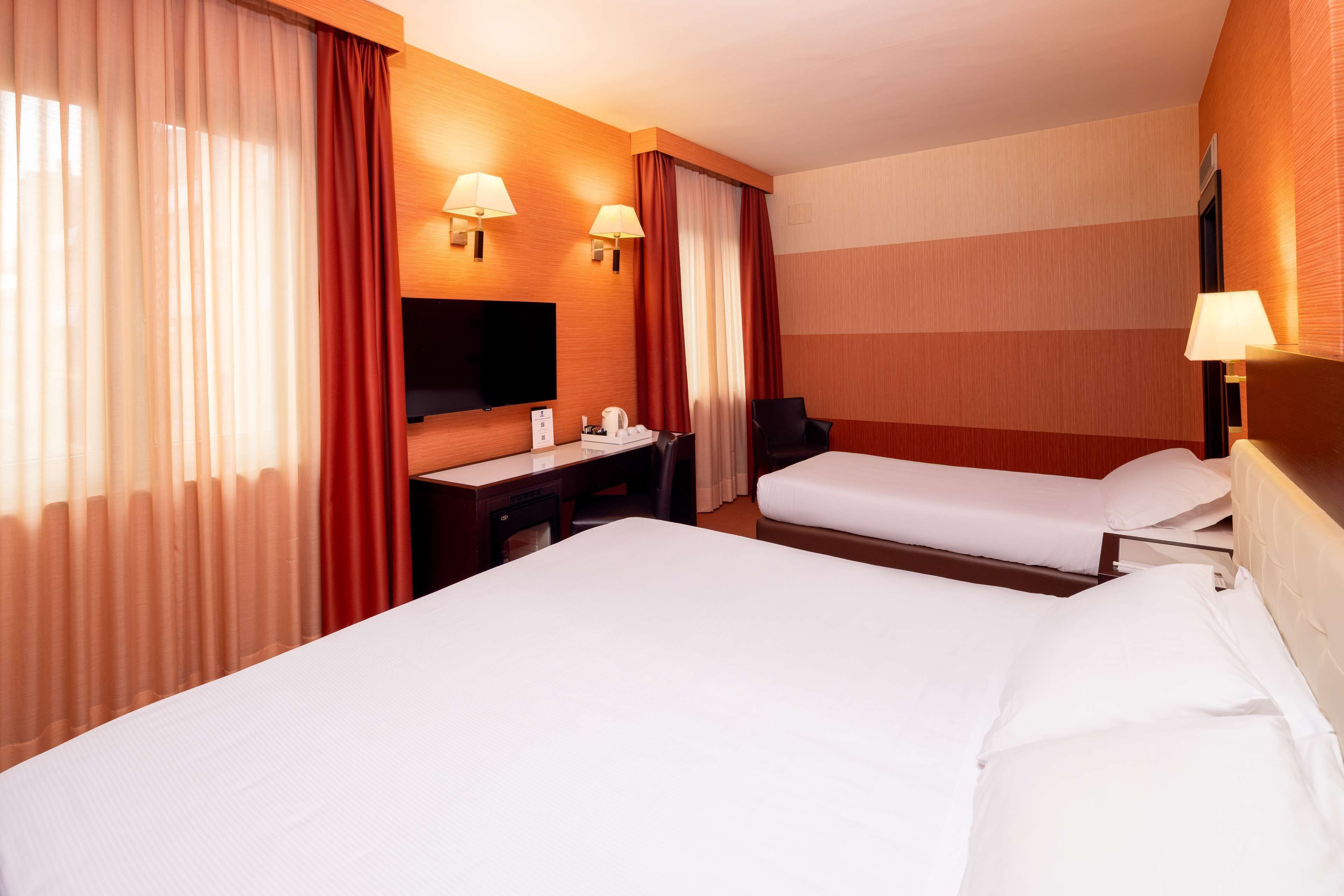A hotel room with two beds, a desk, a chair, a TV, and two wall-mounted lamps.