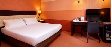 A hotel room with a large bed, a desk with a chair, a television, and two wall-mounted lamps.