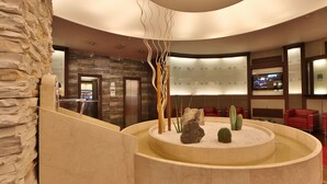 Un hall con pared de piedra, una fuente circular con rocas y cactus, asientos rojos y una pantalla de televisión.