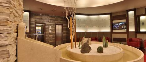 A lobby with a stone wall, a circular water feature with rocks and cacti, red seating, and a television screen.