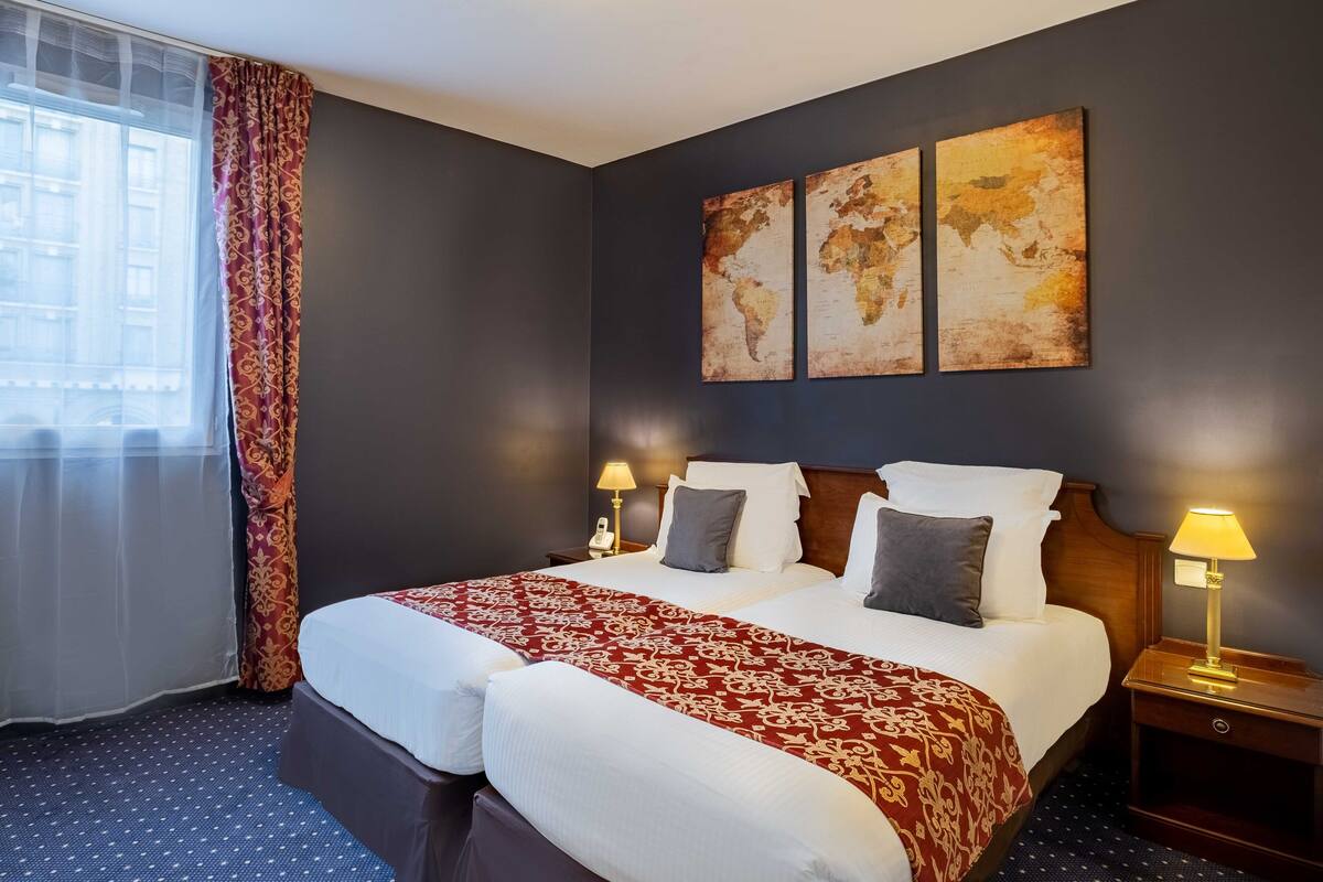 A hotel room with a large bed, two bedside lamps, a world map artwork on the wall, and a window with sheer curtains.