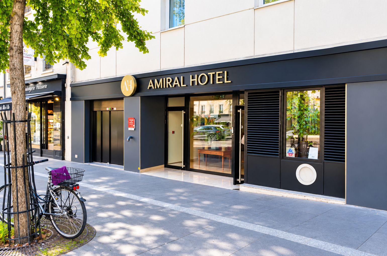 A modern hotel entrance with a glass door, a bicycle parked outside, and a tree providing shade.